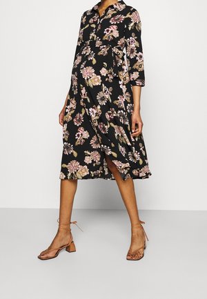 Gravid person iført en sort midi-kjole med blomsterprint med kvartærmede og brune hælede sandaler med ankelremme.