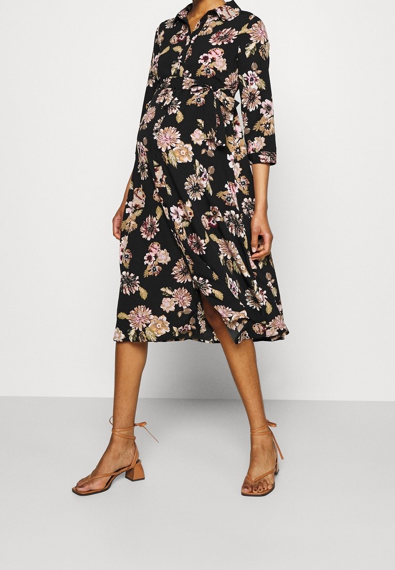 Personne enceinte portant une robe midi noire à fleurs avec des manches trois-quarts et des sandales à talons beiges avec brides à la cheville.