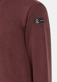 Felpa a maniche lunghe di color marrone scuro con una texture liscia, caratterizzata da un'etichetta nera sulla manica sinistra con accenti bianchi e dorati.