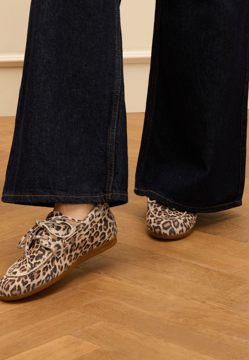 Schuhe im Leopardenmuster mit flacher Sohle und Schnürdesign, kombiniert mit weit geschnittenen, dunklen Jeans auf einem Holzboden.