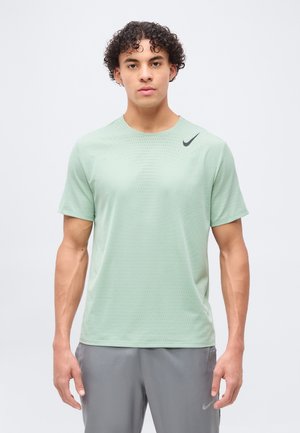 Mladi moški z kodrastimi lasmi, oblečen v svetlo zeleno Nike majico kratkih rokavov in sive Nike hlače, stoji z rokami sproščenimi ob telesu.