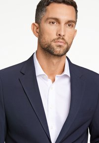 Granatowa marynarka o slim fit i klapach w kształcie notcha, noszona na białej koszuli z kołnierzykiem. Posiada kieszeń na piersi i gładką fakturę materiału.