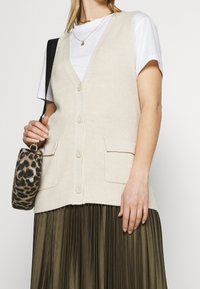 Beige ribbad stickad väst med V-ringning, knappfram och två stora fickor, parat med en vit t-shirt och en veckad olivgrön kjol.