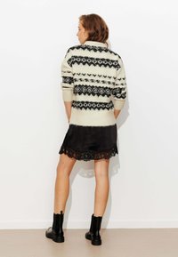 Pull en maille crème avec motifs géométriques noirs, associé à une robe en satin noire ornée d'un ourlet en dentelle, et des bottines noires.