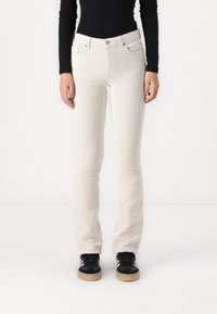 Vita kordflöjlsflarejeans med framfickor och knappstängning, i kombination med svarta skor. Slät yta och vertikalt ribbat mönster.