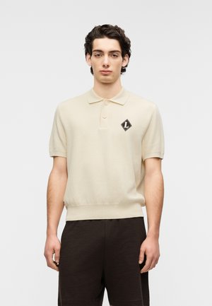 Polo de punto beige con un parche de logotipo en forma de diamante, mangas cortas y cuello estándar. Tejido texturizado con cintura ajustada.