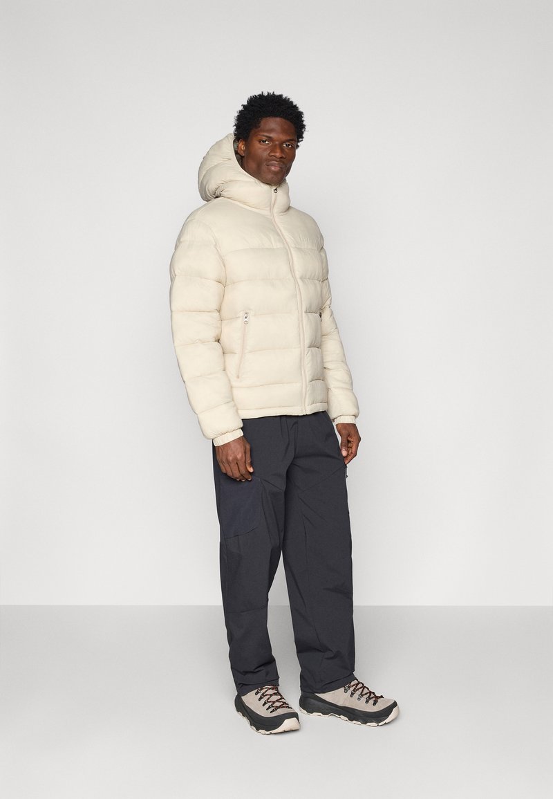 Jasna kremowa kurtka puchowa z kapturem, z zamkiem błyskawicznym z przodu i dwoma bocznymi kieszeniami. Ciemnoszare spodnie cargo i jasne buty trekkingowe.