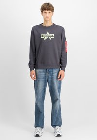 Sötétszürke pulóver csillag grafikával és "INDUSTRIES" felirattal, bordázott szegéllyel és oldalsó cipzáras zsebfedéssel, világoskék farmernadrággal párosítva.