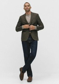 Brun tweedblazer över en ljusgrå tröja, tillsammans med mörkblå jeans och bruna mockaskor, vilket skapar en smart-casual look.