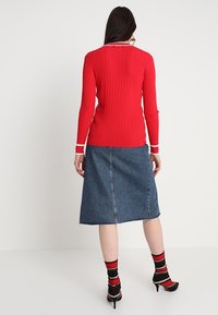 Haut à manches longues rouge côtelé avec des bordures blanches, associé à une jupe en denim bleu. Le mannequin porte des talons noirs et des chaussettes rayées rouges et noires.