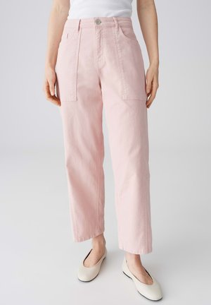 Vrouw draagt hoge taille, wijde lichtroze broek met voorzakken, witte mouwloze top en crème ballerina's.