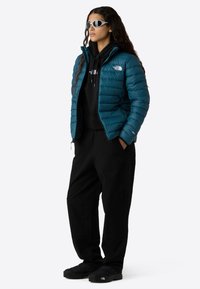 Donna che indossa un piumino teal, una felpa nera con cappuccio, pantaloni neri, scarpe nere e occhiali da sole bianchi avvolgenti, in piedi con le mani nelle tasche.
