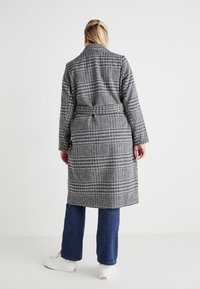 Γκρι καρό παλτό με ζώνη στη μέση, μακριά μανίκια και μεσαίου μήκους κόψιμο, σε συνδυασμό με μπλε τζιν και λευκά αθλητικά παπούτσια.