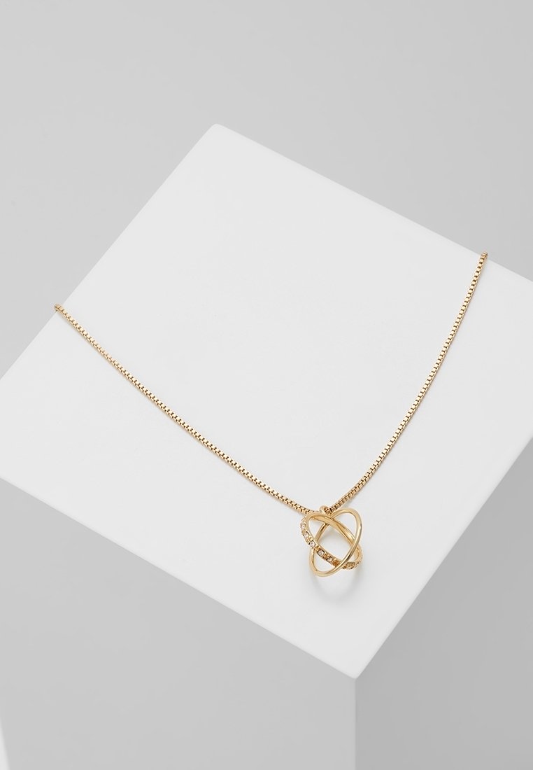 Collier en chaîne d'or délicat avec pendentif en cercle entrelacé orné de petits cristaux intégrés, présenté sur une surface géométrique blanche.