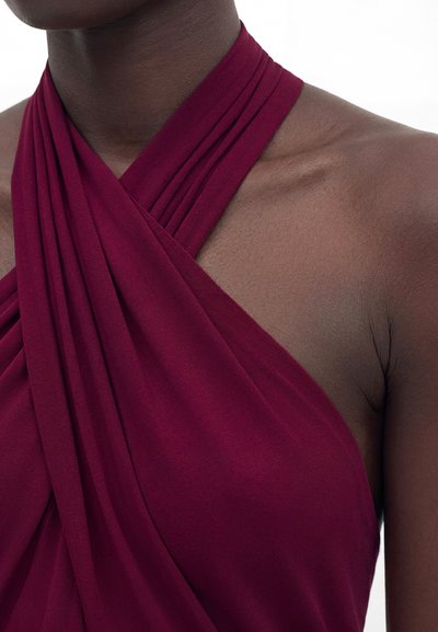 Άτομο με σκούρο δέρμα που φοράει ένα αμάνικο, βαθύ κόκκινο τοπ με ύφασμα που διασταυρώνεται στο ντεκολτέ και στον ώμο.