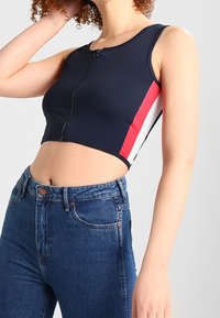 Top corto navy con zip frontale, caratterizzato da strisce laterali rosse e bianche. Abbinato a jeans in denim blu a vita alta.