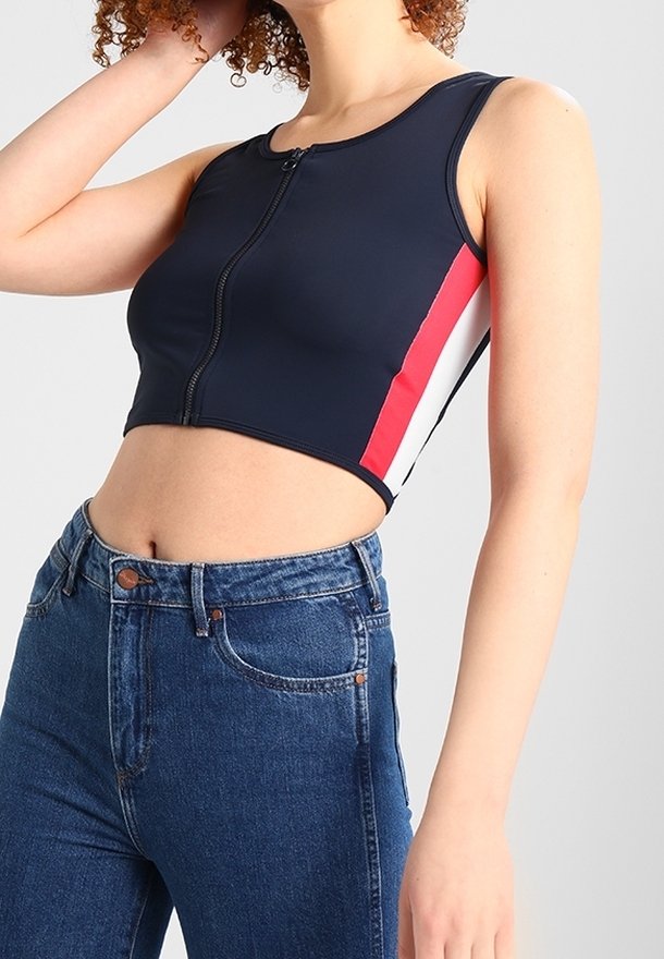 Top corto navy con zip frontale, caratterizzato da strisce laterali rosse e bianche. Abbinato a jeans in denim blu a vita alta.