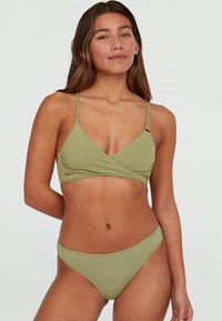 Kobieta z długimi brązowymi włosami, ubrana w oliwkowozielony bikini z górą w stylu owiniętym i dopasowanymi majtkami, na jednolitym jasnym tle.