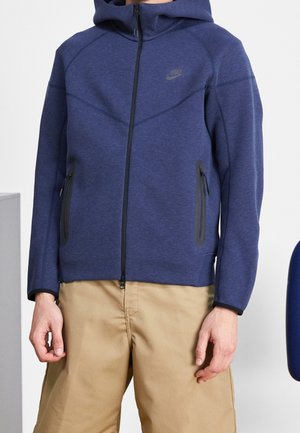 Man draagt een marineblauwe Nike hoodie met rits, met zwarte rits en zakdetails, gecombineerd met lichtbruine broek.