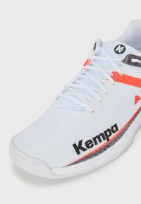 Hvite trening sko med mesh-overdel, med oransje og grå detaljer, flate snøringer og en svart "Kempa" logo på siden.