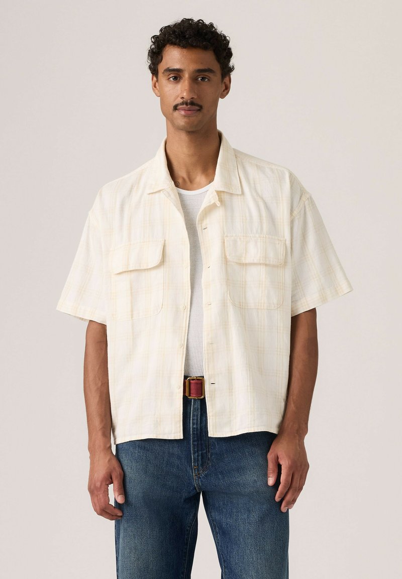 Mann mit lockigem Haar und Schnurrbart, der ein beiges kurzärmeliges Hemd über einem weißen Tanktop und blauen Jeans vor einfachem Hintergrund trägt.