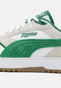 Zapatillas Puma con ante beige y acentos texturizados verdes, superposiciones sintéticas blancas y cordones blancos, con una suela de goma marrón.