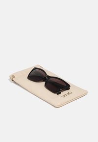 Occhiali da sole oversize neri su una morbida custodia beige con logo dorato. Gli occhiali hanno lenti scure e una montatura spessa con angoli arrotondati.