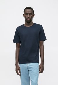 Uomo con una maglietta a maniche corte blu navy e jeans azzurri, in piedi di fronte a uno sfondo bianco semplice, rivolto verso l'obbiettivo.