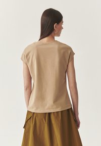 Beige, rövid ujjú felső, kerek nyakkivágással és laza szabással. Puha tapintás, egyszínű dizájn és varrott szegély. Olívazöld szoknyával viselve.