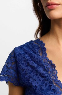 Top in pizzo blu navy con motivi floreali, scollatura a scallop e maniche corte. Il tessuto testurizzato crea un contrasto con il tono della pelle liscia.