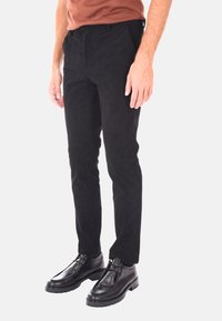 Uomo vestito con pantaloni in velluto a coste neri e scarpe eleganti in pelle nera, in piedi contro uno sfondo chiaro e uniforme.
