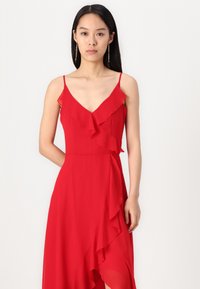 Crvena chiffon haljina s V-izrezom, tankim naramenicama i slojevitim ruffles uz prednji dio i rub, stvarajući asimetričan oblik.