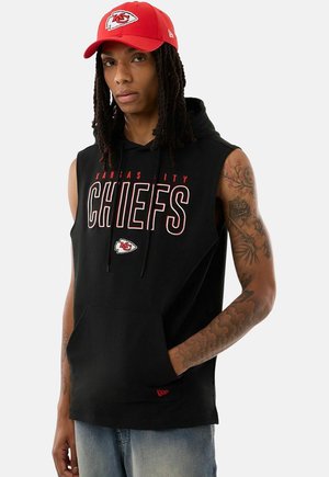 Person mit langen Dreadlocks, die eine rote Kansas City Chiefs Kappe und ein ärmelloses schwarzes Kapuzensweatshirt mit Teamnamen und Logo trägt, Hände in den Taschen.