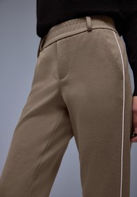 Beige taps toelopende broek gemaakt van gestructureerde stof, met een elastische tailleband en witte zijstrepen. Twee zijzakken zichtbaar.