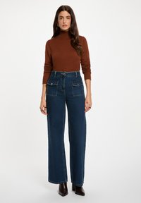 Jean en denim à taille haute et jambes larges de couleur foncée, avec des poches plaquées à l'avant et des finitions en métal argenté. Assorti à un pull col roulé ajusté de couleur marron.