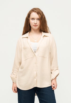 Junge Frau mit langen roten Haaren, die ein hellbeiges Hemd über einem weißen Top und dunklen blauen Jeans trägt, steht vor einem weißen Hintergrund.