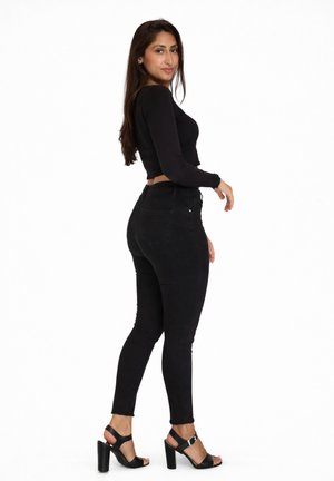 Kvinde med langt mørkt hår iført en tætsiddende sort langærmet top, sorte skinny jeans og sorte højhælede sandaler, stående i profil.