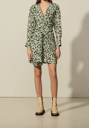 Vrouw draagt een groene wikkeljurk met madeliefjesprint en lange mouwen, gecombineerd met beige enkellaarzen met zwarte zolen.