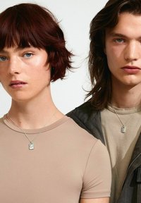 Dos personas llevan collares de plata con colgantes rectangulares. La mujer viste una blusa ajustada beige, y el hombre lleva una camisa beige clara debajo de una chaqueta oscura.