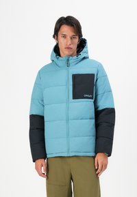 Uomo che indossa un giubbotto invernale Oakley blu e nero con cappuccio e pantaloni verde oliva, in piedi su uno sfondo bianco.