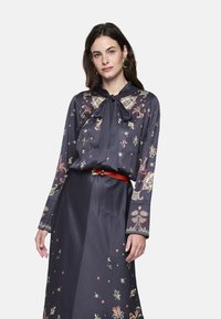 Marineblaue Bluse mit Blumenmuster und Bindeband am Hals, kombiniert mit einem passenden langen Rock. Mit rotem Gürtel und dekorativen Akzenten in Gold. Glatte Textur.