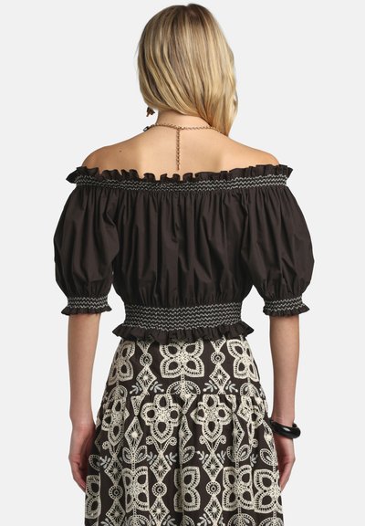 Femme blonde portant un haut noir à épaules dénudées avec des manches ballon et une jupe noire et blanche à motifs, vue de dos.
