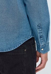 Denim shirt in light blue with long sleeves. Features a rounded hem, buttoned cuffs, and contrasting stitching on a dark fabric background.