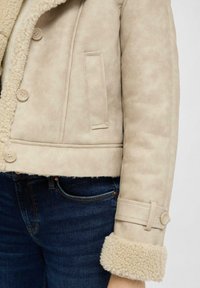 Beige shearling-gewatteerde jas met knoopsluiting en riem_detail op mouwen, gedragen met donkerblauwe spijkerbroek.