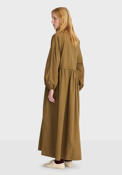 Femme aux longs cheveux blonds portant une robe longue et ample couleur olive-brun et des chaussures crème, debout de profil devant un fond clair uni.