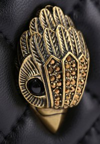 Złoty dekoracyjny klips z motywem pióra, ozdobiony małymi brązowymi cyrkoniami i czarnym kamieniem szlachetnym, umieszczony na czarnym materiałowym tle o fakturze.