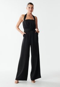 Woman wearing a black halter-neck jumpsuit with wide legs, a waist tie, hands in pockets, and gold high-heeled sandals against a white background.