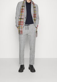 Šedý strukturovaný oblek s černým rolákem, doplněný kostičkovaným šátkem v červené, žluté a šedé barvě. Černé boty dotvářejí celkový vzhled.