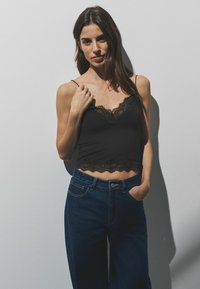 Melns kamzolis ar mežģīņu apmali un plānām lencītēm, kombinēts ar augsti jostasvietas ziliem džinsa biksēm. Gluda auduma, pieguļošs dizains ar viļņotām malām.