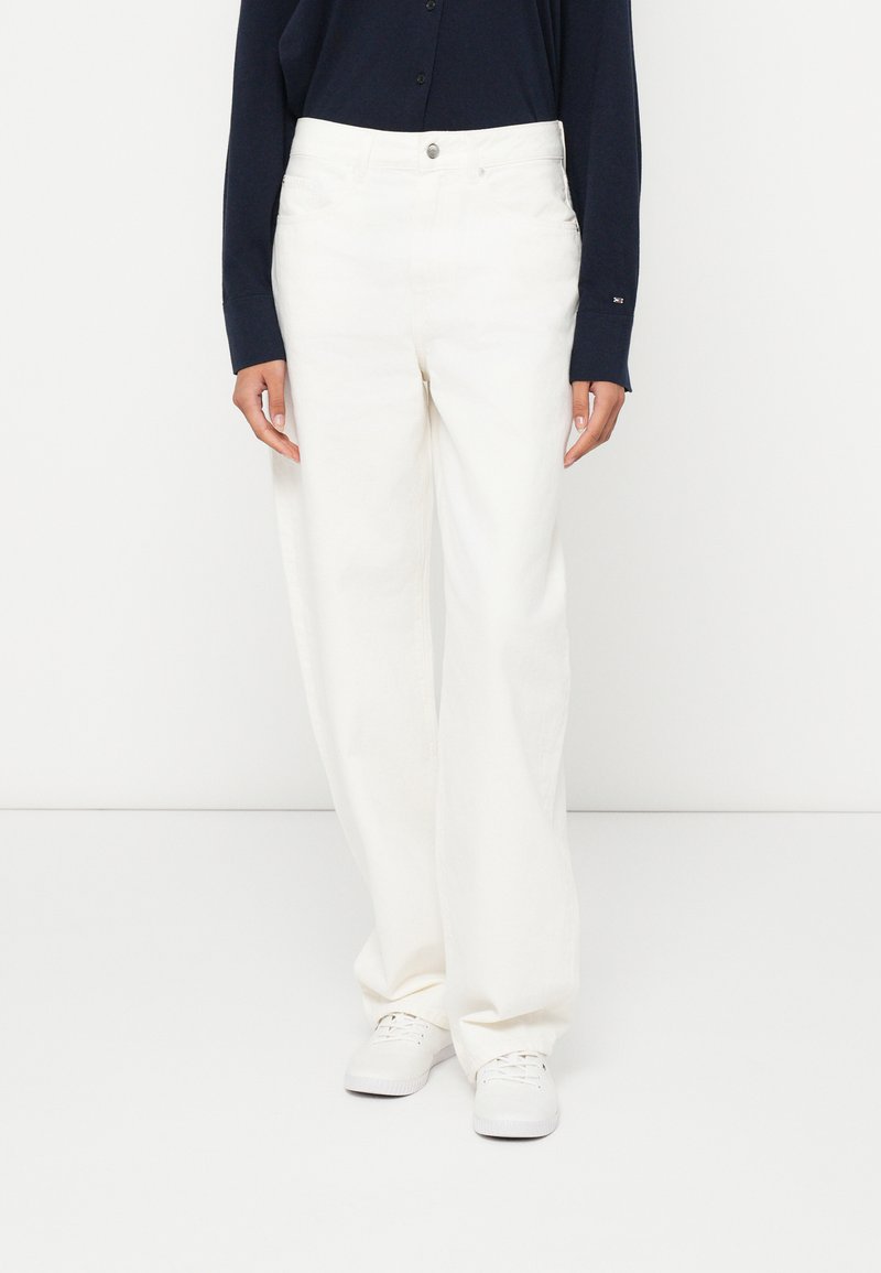 Biele nohavice s širokými nohavicami a vysokým pásom, vyrobené z hladkej látky, s predným zapínaním na gombíky a bočnými vreckami. Nosia sa s bielou obuvou.